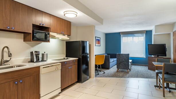 A modern kitchen with a dishwasher, microwave, and refrigerator. There is a dining area with a table and chairs, and a TV in the background.