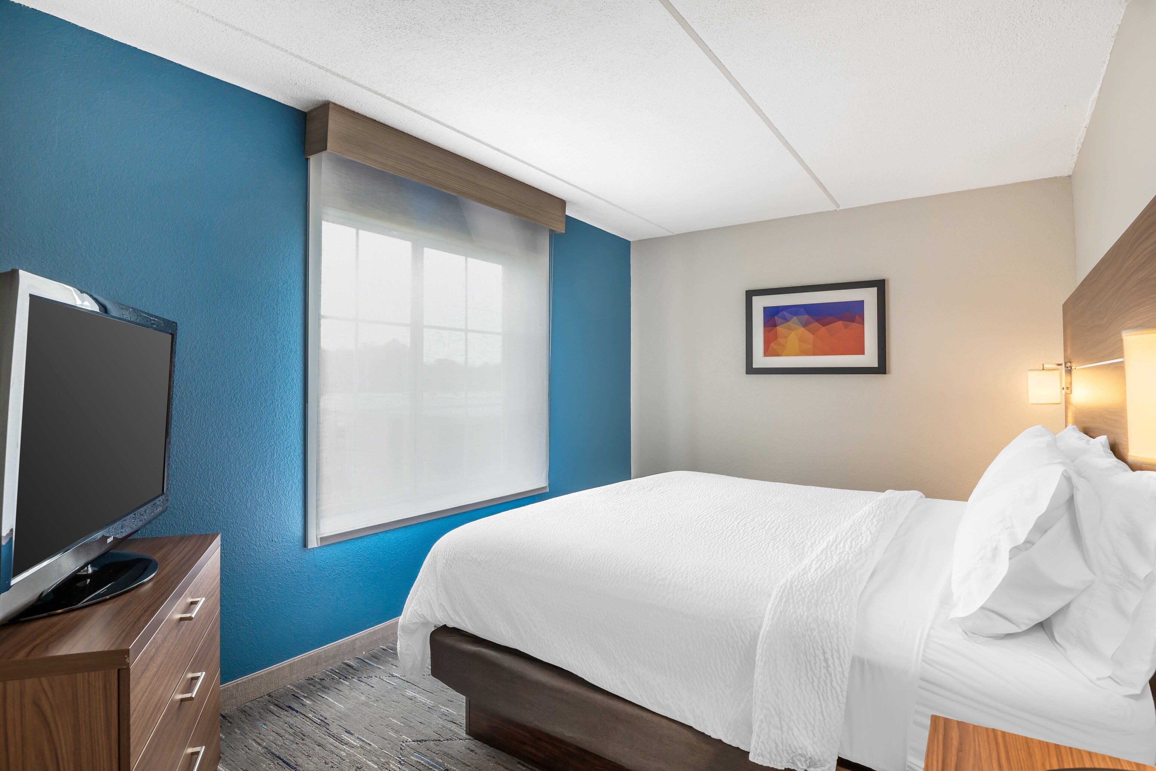 Habitación de hotel con una cama, un televisor en un aparador de madera, una ventana con persianas y un cuadro en la pared.