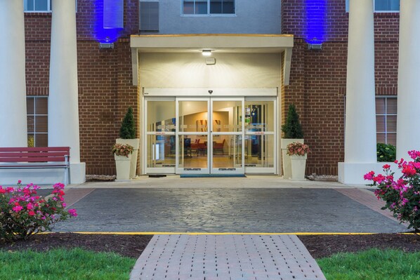 A building entrance with a glass door, a brick facade, and a paved walkway leading to a bench.