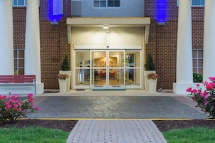 A building entrance with a glass door, a brick facade, and a paved walkway leading to a bench.