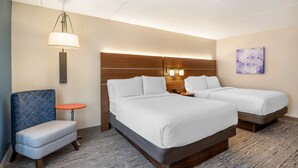 A hotel room with two beds, a small table, a chair, and a wall-mounted lamp.