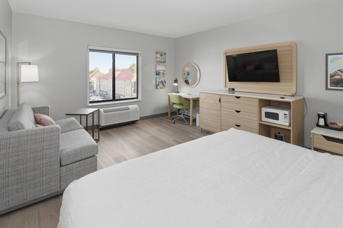 A modern hotel room with a large bed, a desk, a chair, a flat-screen TV, a microwave, and a window with a view of buildings.
