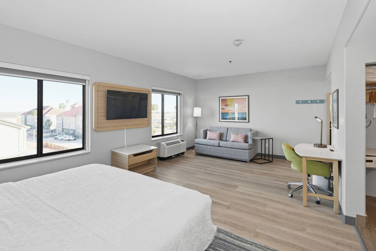 A modern hotel room with a large bed, a desk with a chair, a sofa, and a TV.