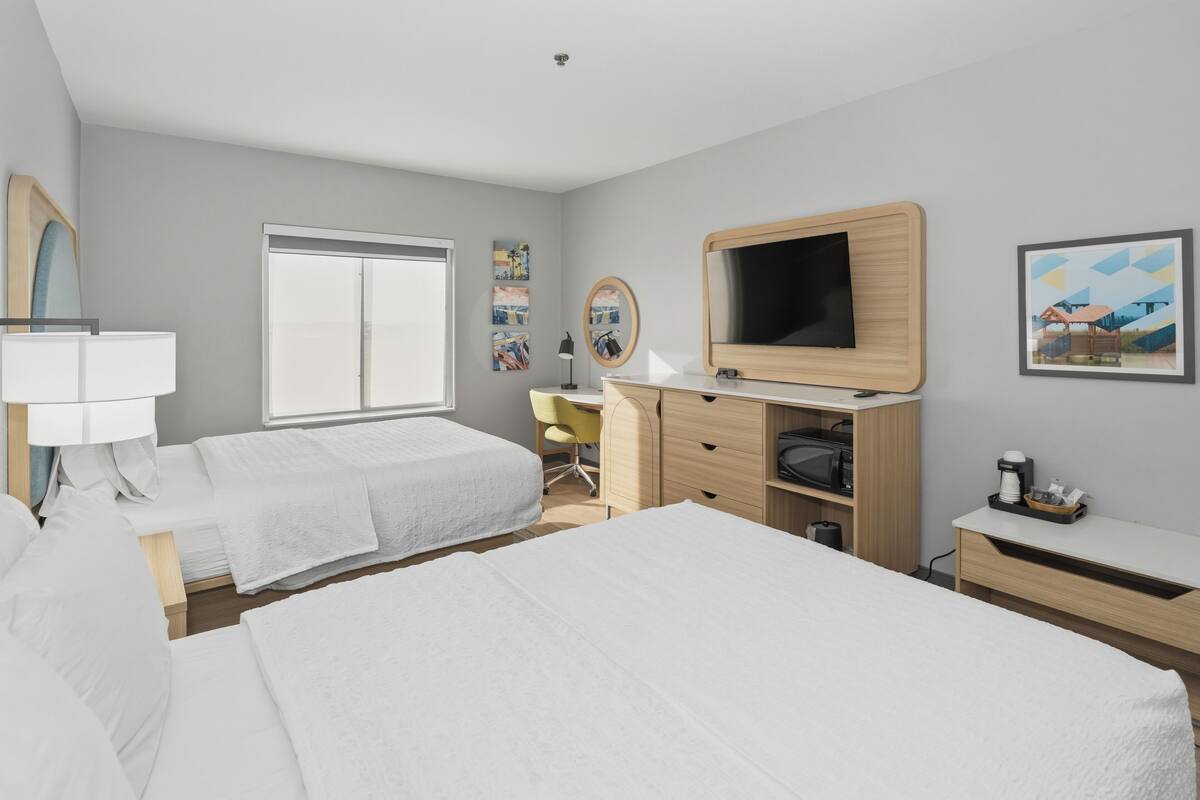 A hotel room with two beds, a desk, a TV, a mirror, and artwork on the walls.