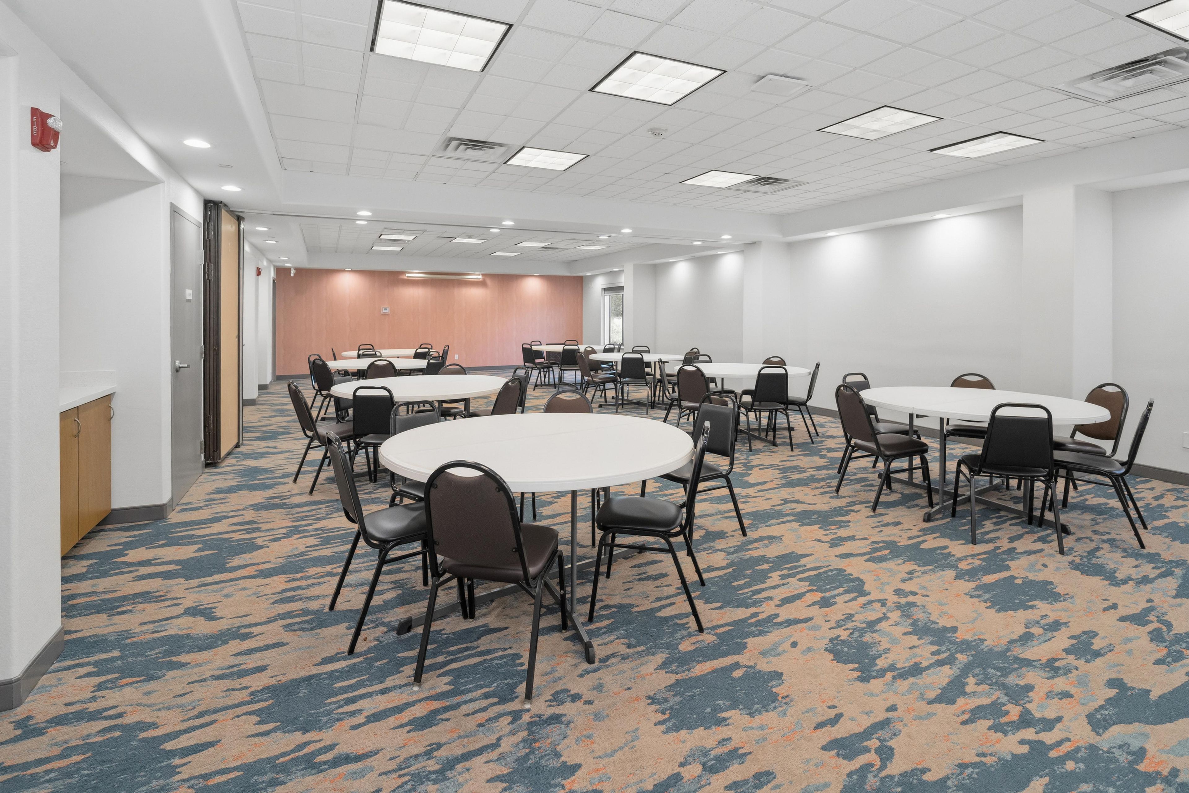 A conference room with multiple tables and chairs, carpeted flooring, and a white ceiling with recessed lighting.