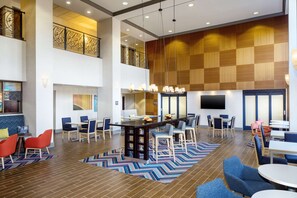 A spacious lobby with a high ceiling, a central bar area, multiple seating arrangements, and a large screen on the wall.