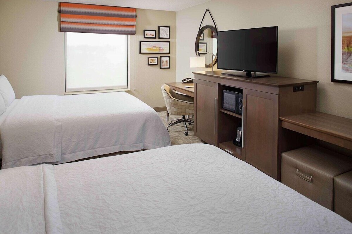 Une chambre d’hôtel avec deux lits, un bureau, une télévision et une fenêtre avec des stores.