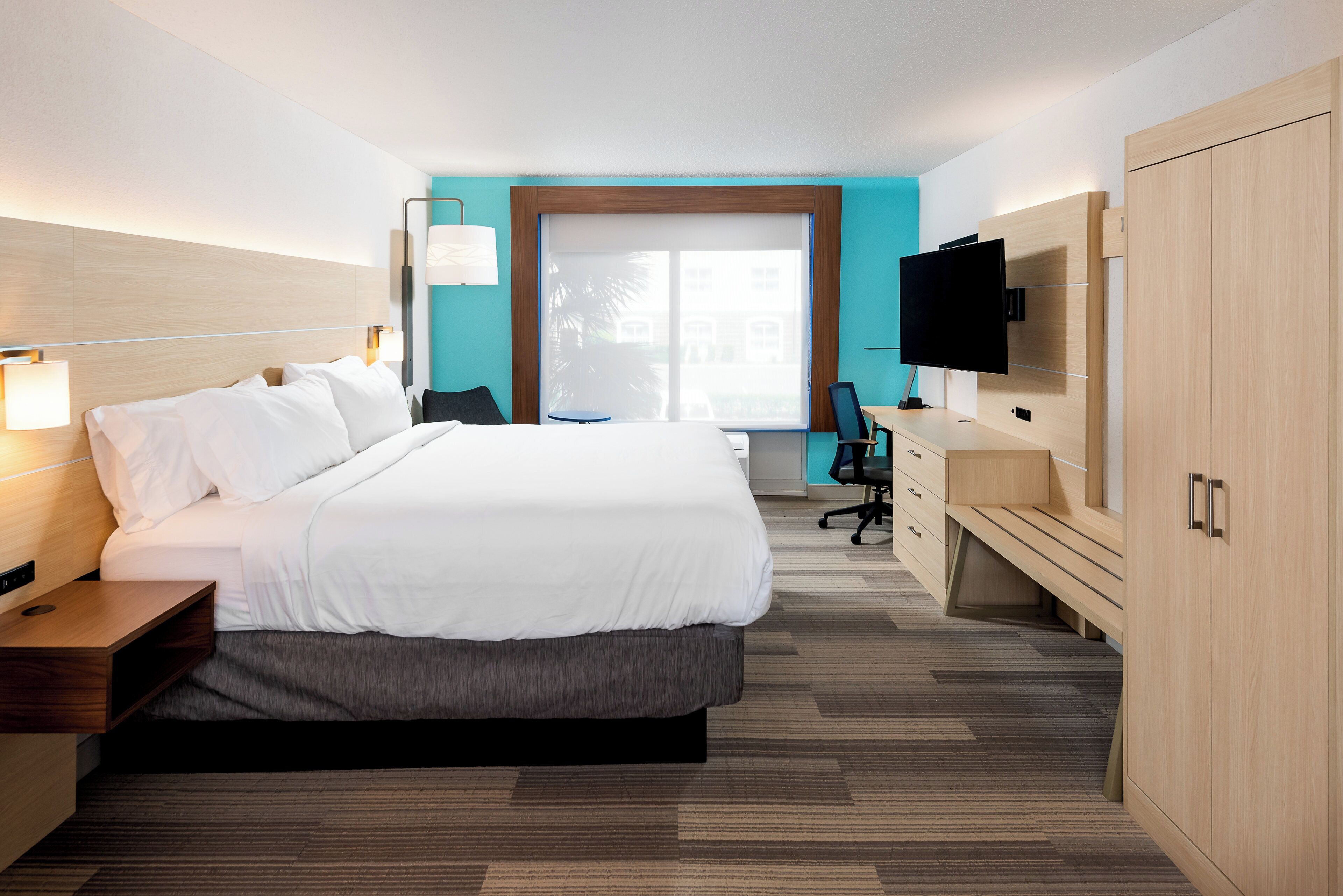 A hotel room with a bed, bedside table, desk, chair, TV, wardrobe, and a window with a view.