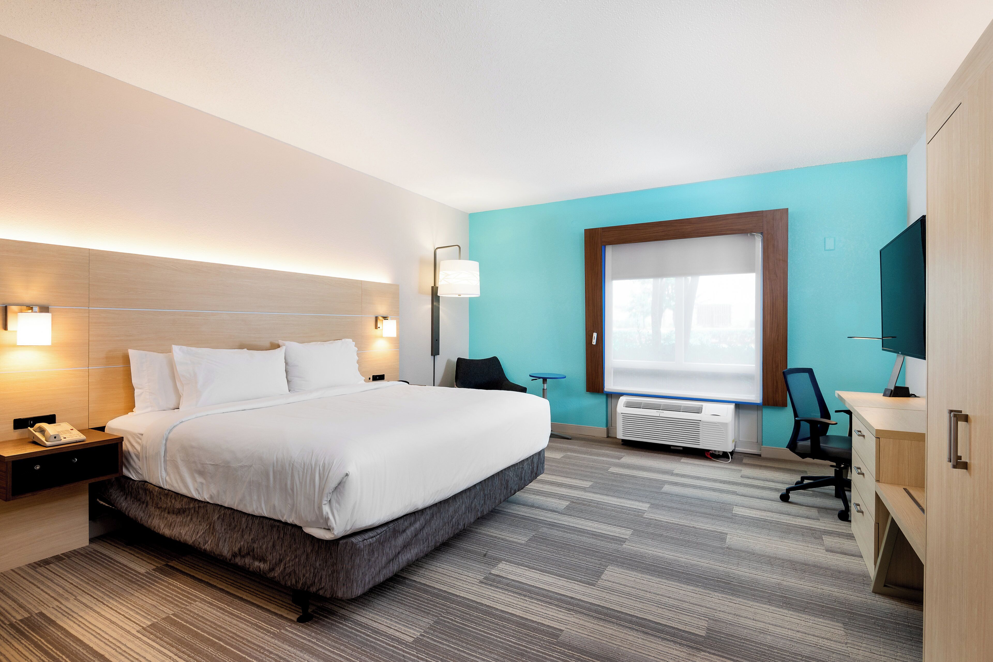 A hotel room with a large bed, bedside lamps, a desk with a chair, a TV, and a window with blinds.
