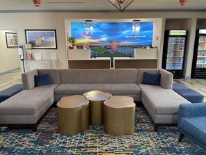 A modern lobby with a sectional sofa, three gold side tables, a large TV screen, and vending machines. - La Quinta Inn & Suites by Wyndham Oklahoma City - Moore (Moore)