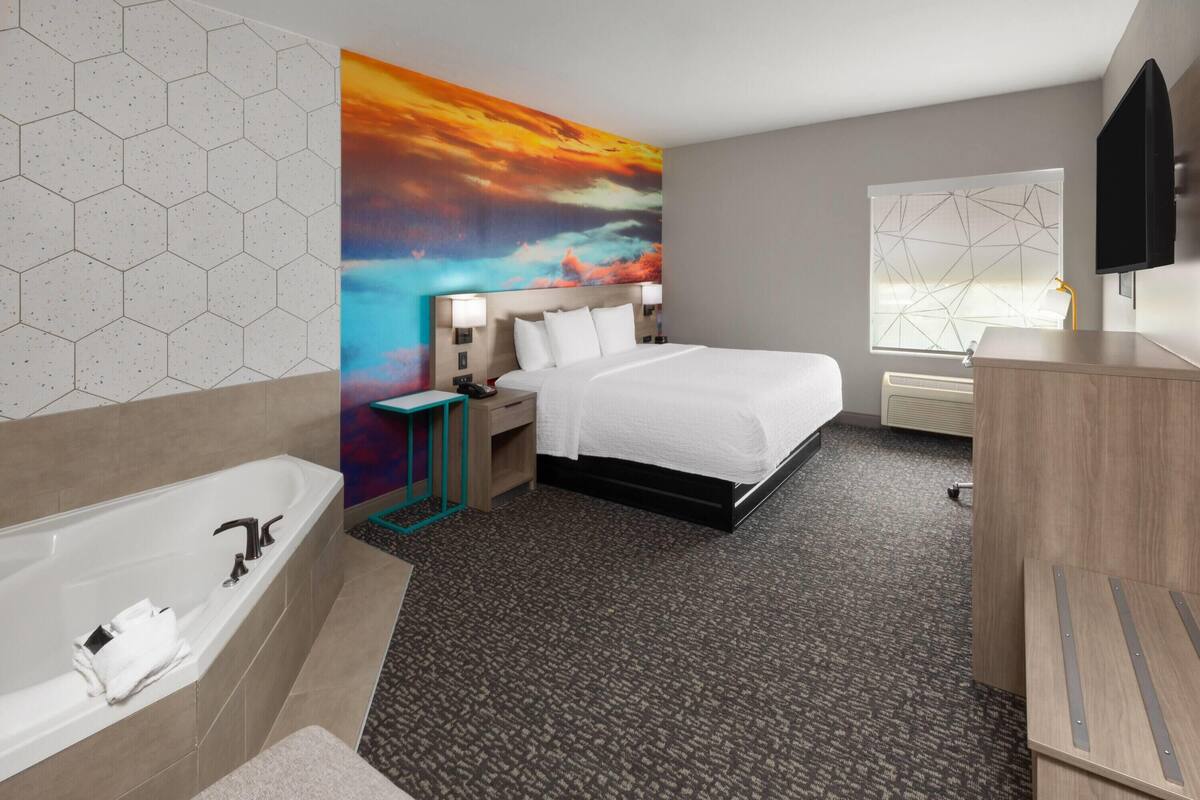 A modern hotel room with a large bed, a bathtub, a TV, and a geometric-patterned window.