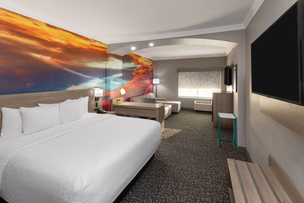 A hotel room with a large bed, a desk, a chair, a TV, and a painting of a sunset on the wall.