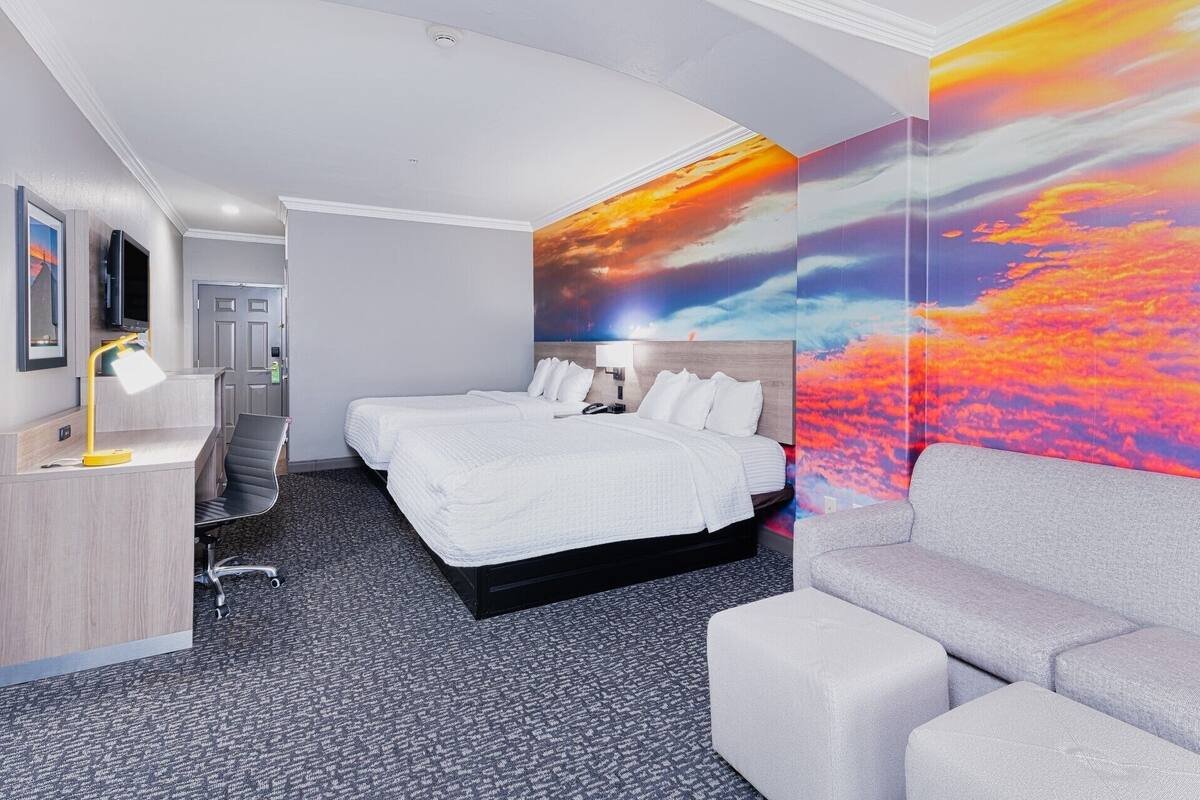 A hotel room with a large bed, a desk with a lamp, a grey sofa, and a vibrant sunset mural on the wall.