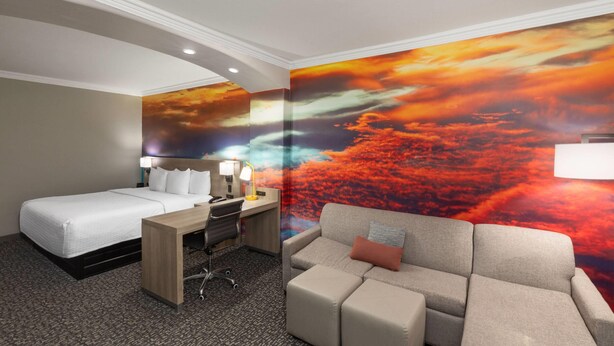 Una habitación de hotel con una cama, un escritorio con una silla, un sofá y un mural grande con un atardecer.