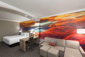 Una habitación de hotel con una cama, un escritorio con una silla, un sofá y un mural grande con un atardecer.