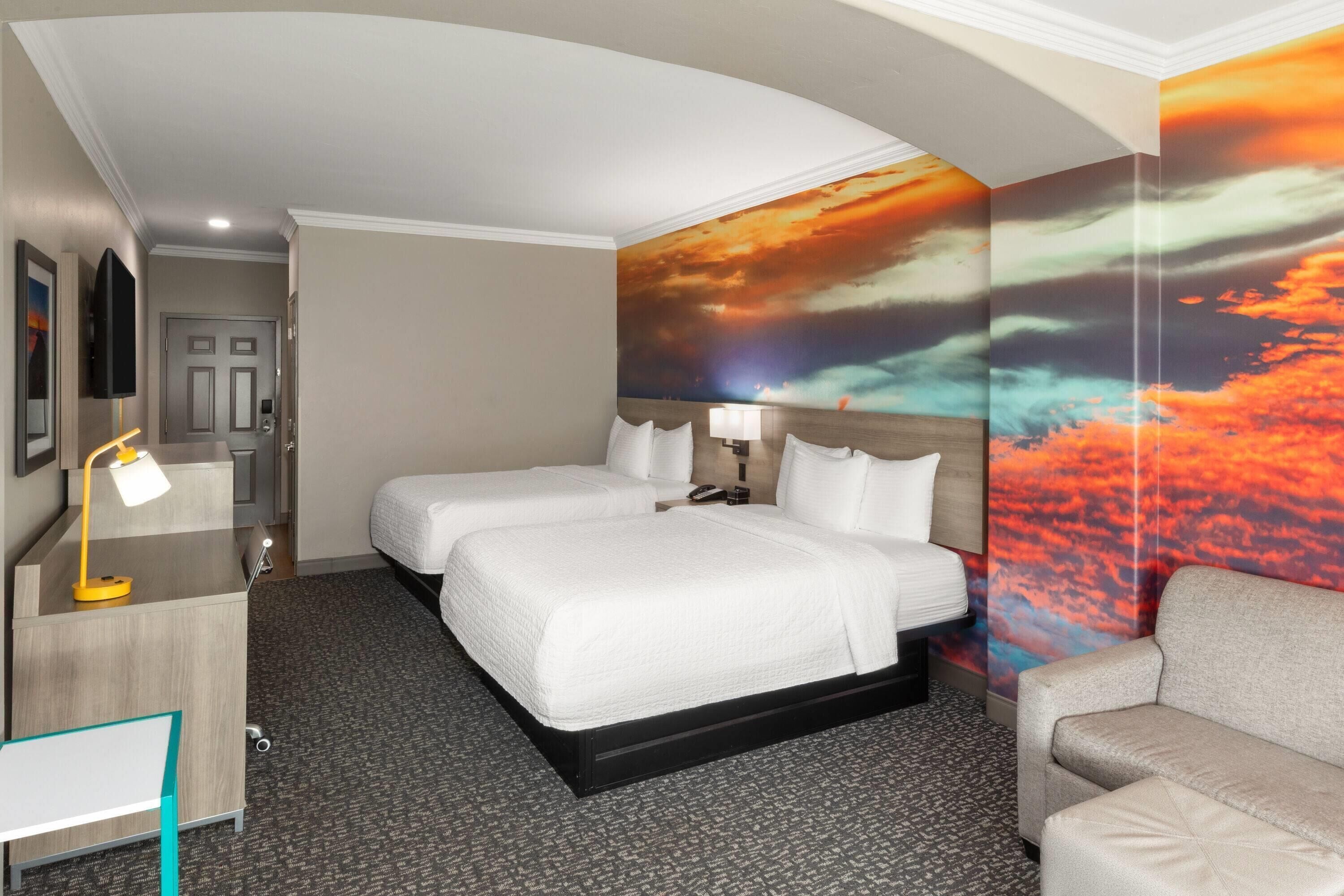 A hotel room with two beds, a desk with a lamp, a sofa, and a large wall mural.