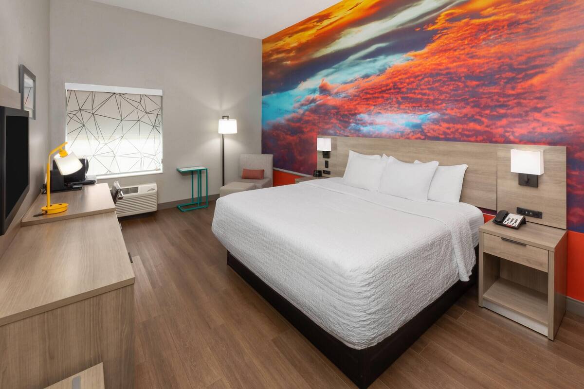 A modern hotel room with a large bed, bedside tables, a desk with a lamp, a large window with geometric blinds, and a vibrant sunset mural on the wall.
