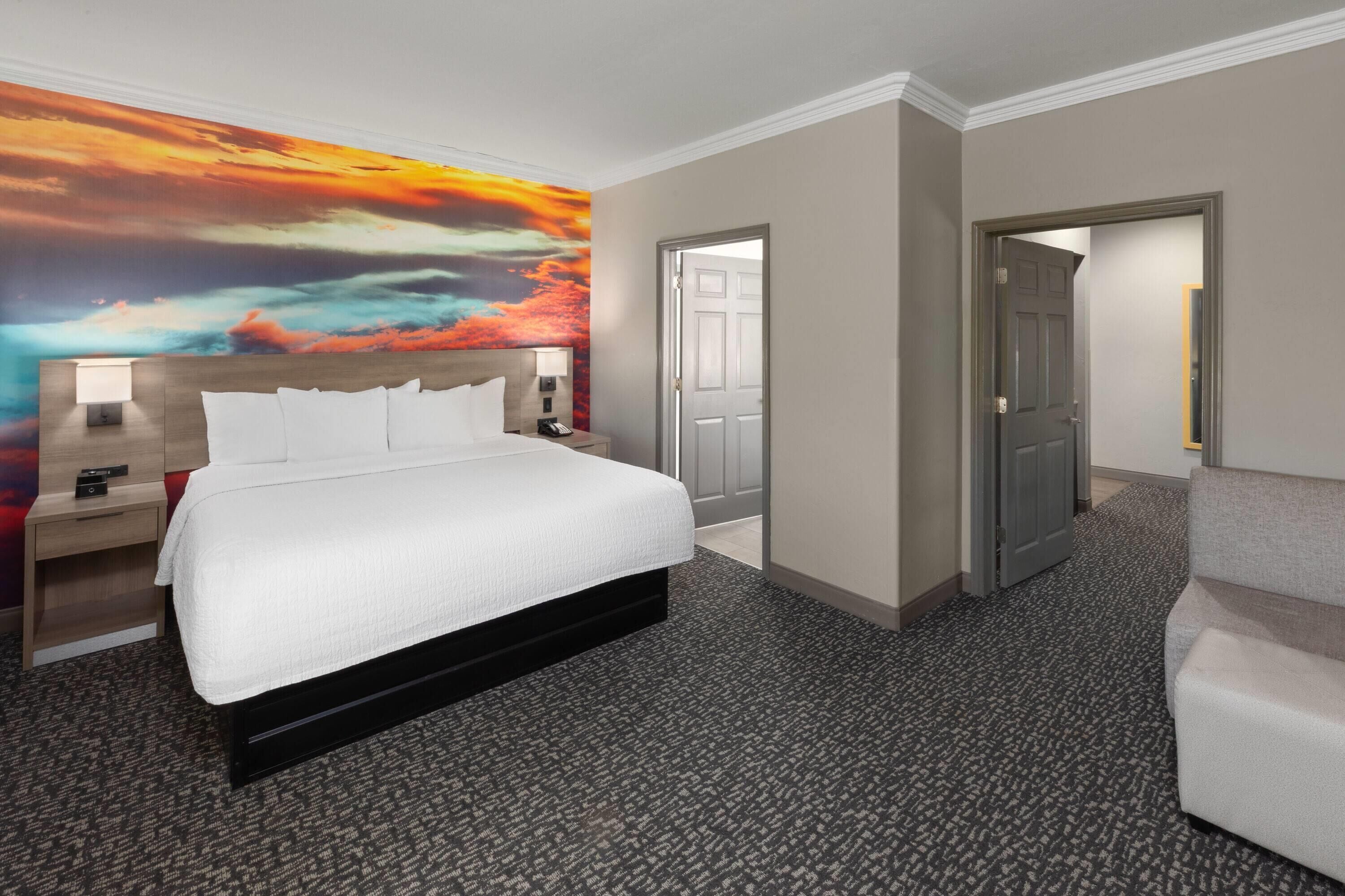 A hotel room with a large bed, a nightstand, a desk, a chair, and a door leading to another room.