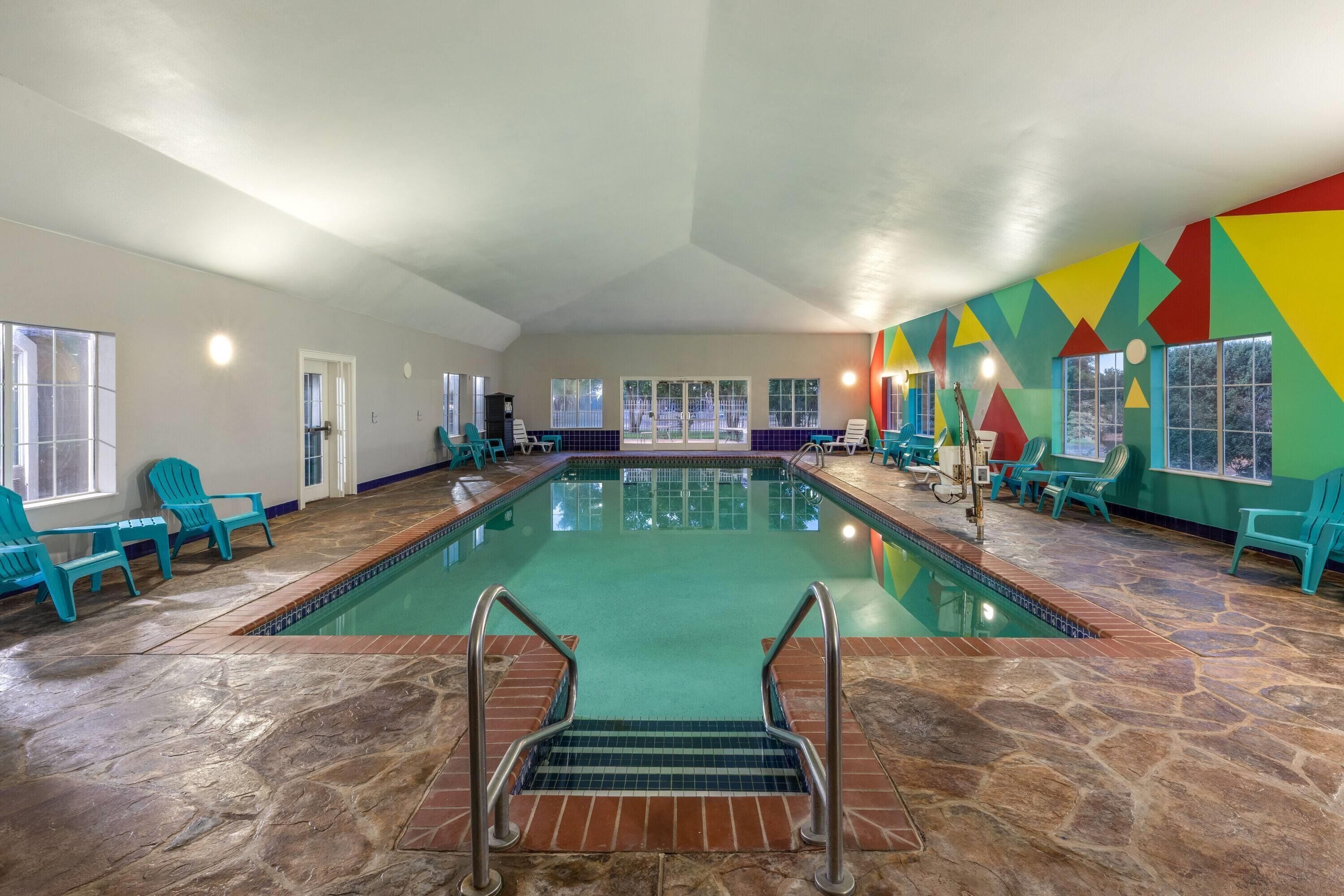 Una piscina interior con agua cristalina, rodeada de sillas turquesa y un llamativo diseño de pared geométrico.