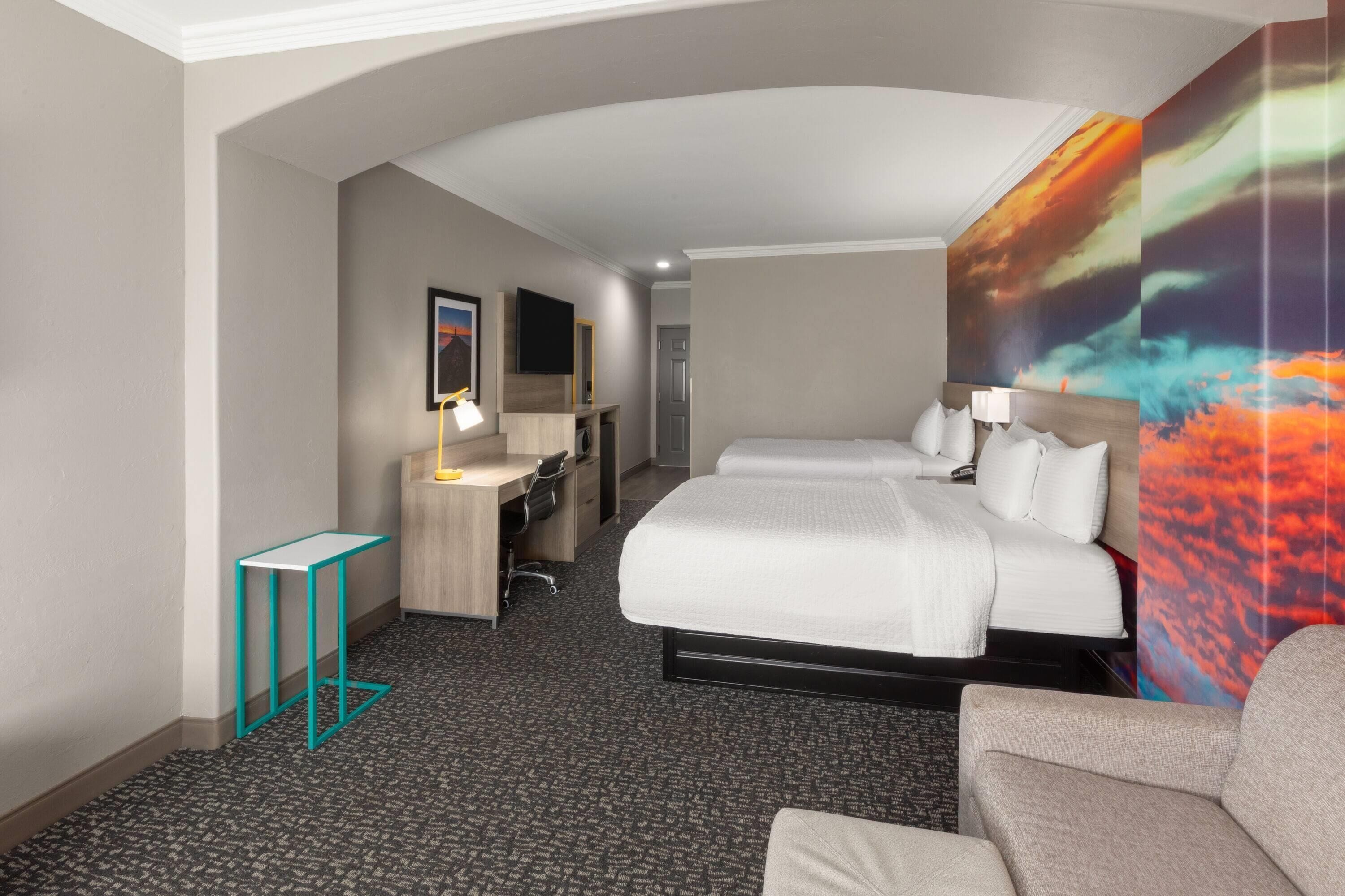 Una habitación de hotel con una cama grande, un escritorio con una silla, una mesita pequeña y un mural grande en la pared.