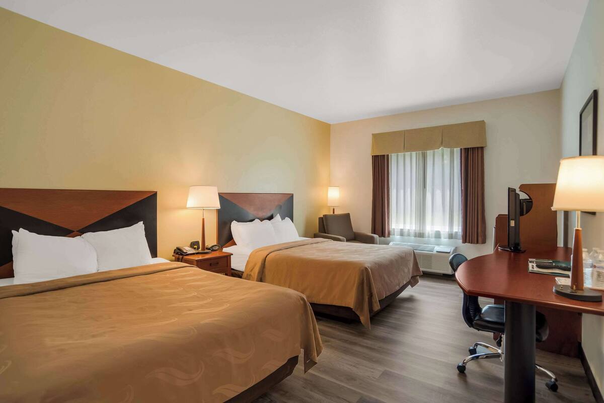 A hotel room with two beds, a desk, and a chair. There are lamps on the desk and nightstands beside the beds.