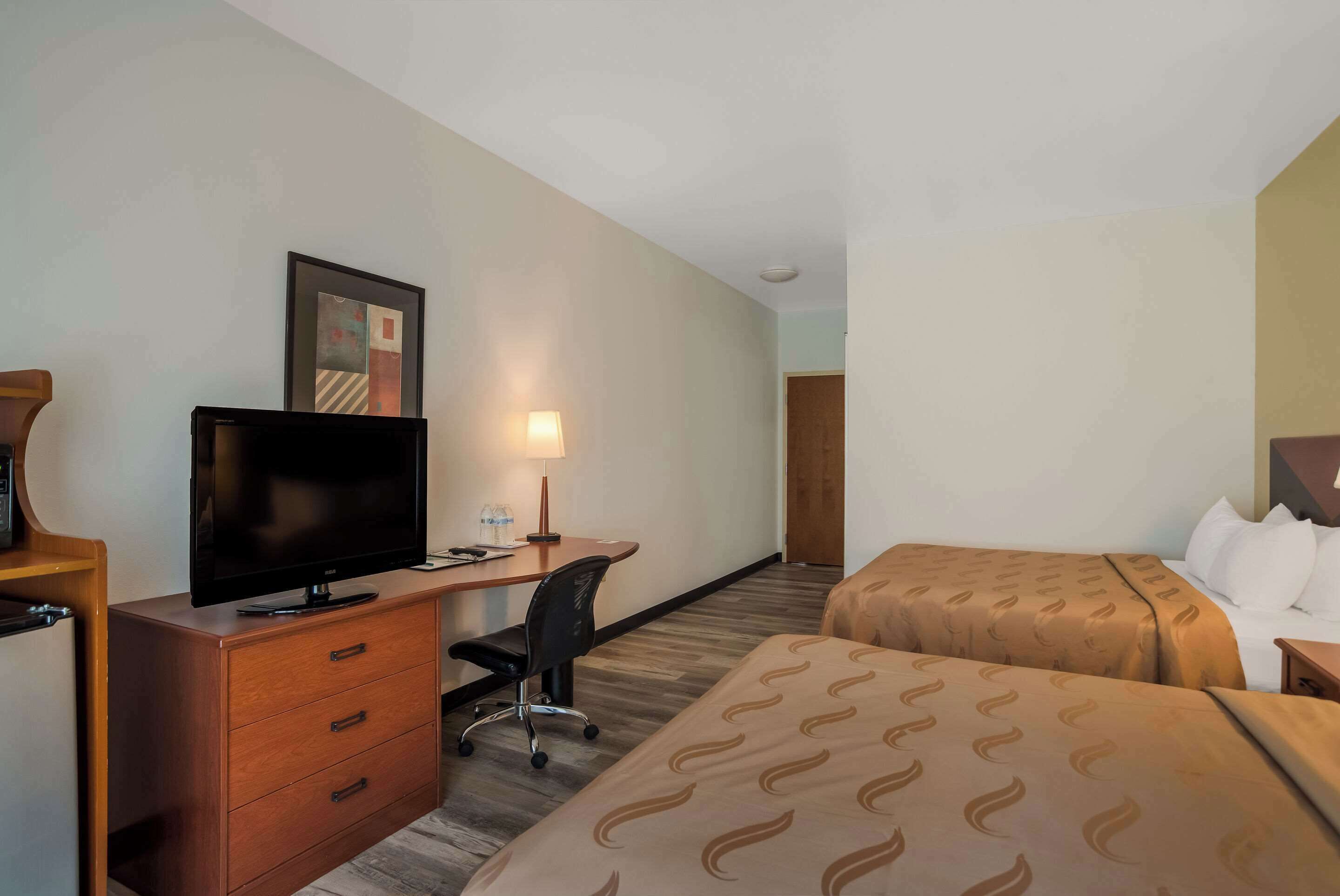 A hotel room with a flat-screen TV, a desk with a chair, a bed with patterned bedding, and a wooden dresser.