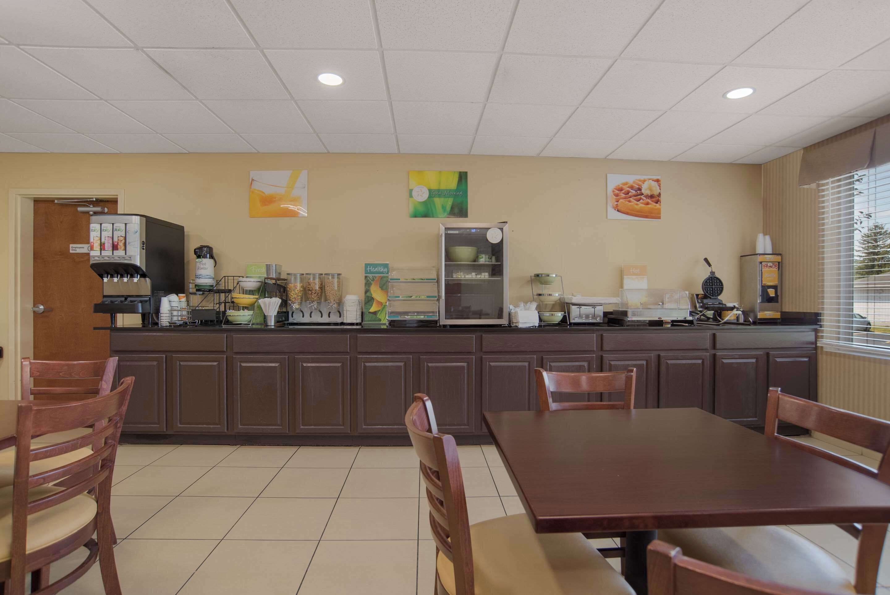 A clean, well-lit kitchen area with a coffee machine, refrigerator, and various kitchen appliances. There are wooden chairs and a table in the foreground, and a window with blinds on the right side.