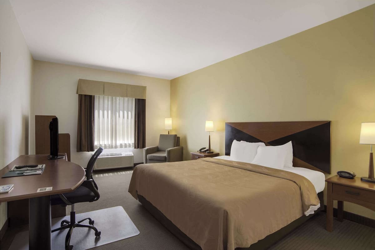 A hotel room with a large bed, a desk, a chair, a desk lamp, a bedside table, and a window with curtains.