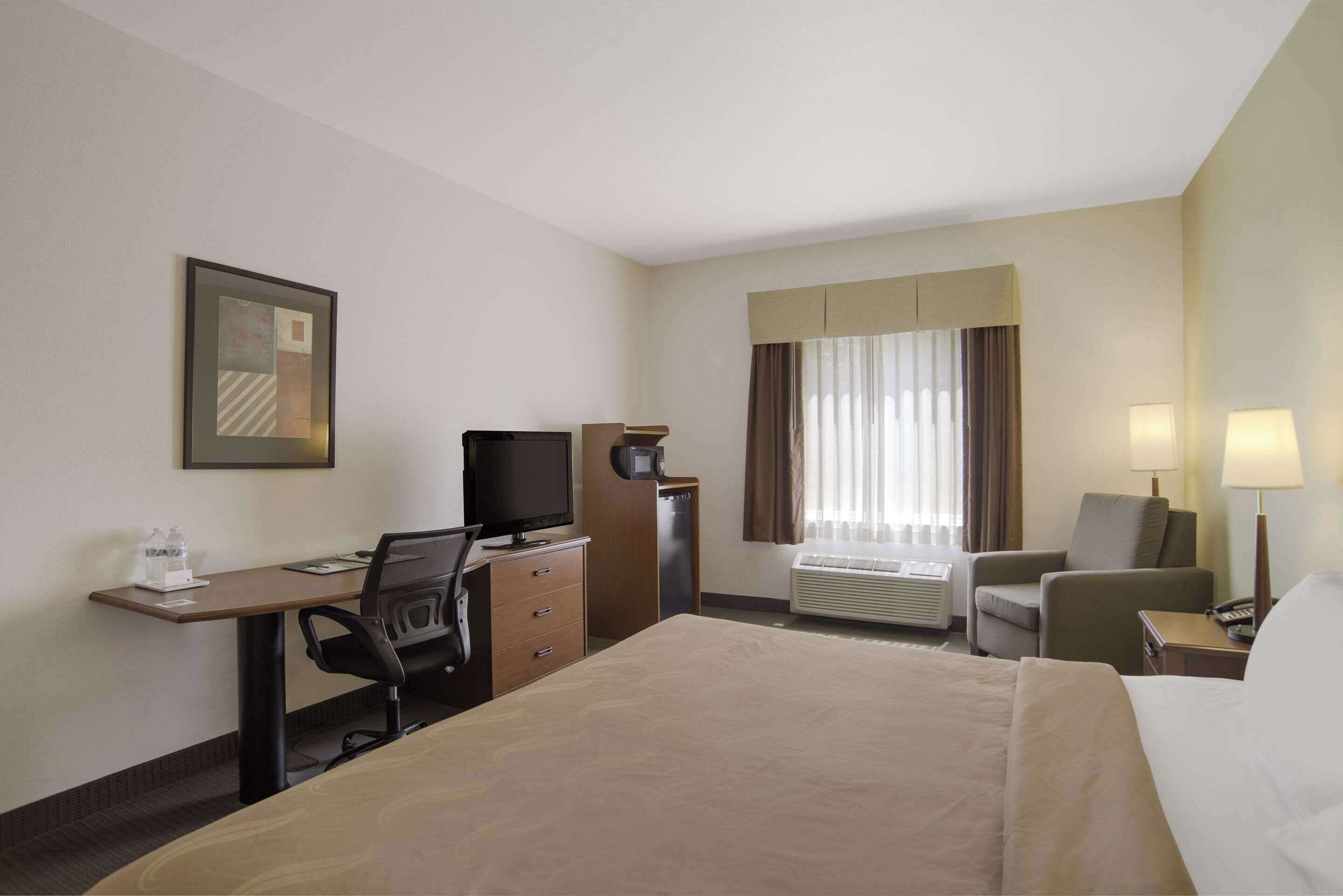 A hotel room with a bed, desk, chair, television, and air conditioning unit.