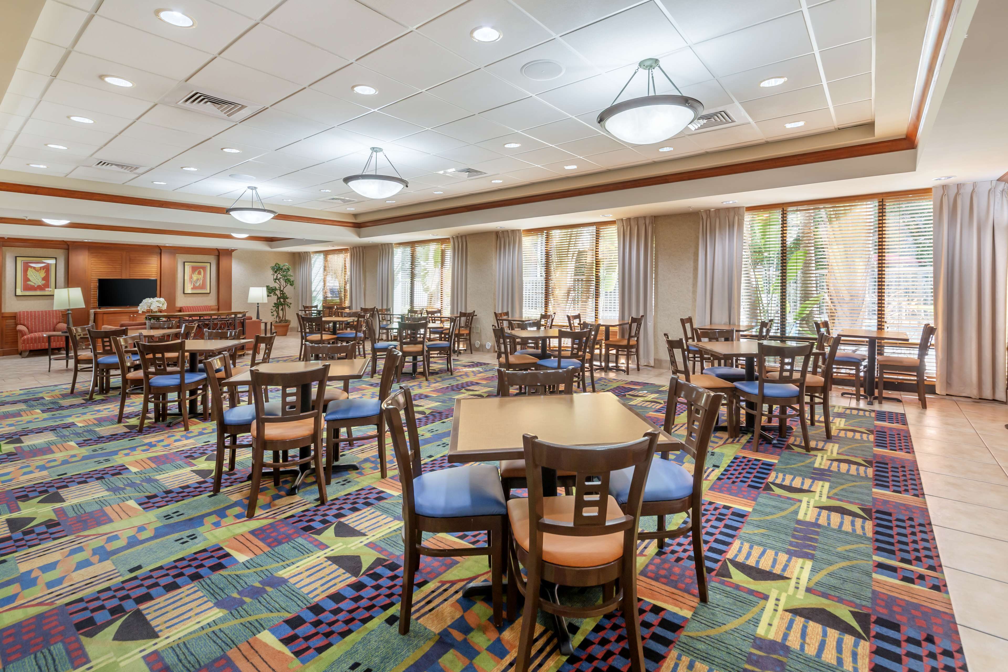 Une salle à manger d’hôtel avec de nombreuses tables et chaises, une moquette à motifs et de grandes fenêtres avec des rideaux.