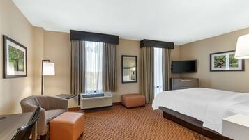 Habitación de hotel con cama, escritorio, silla, reposapiés, televisor y espejo.