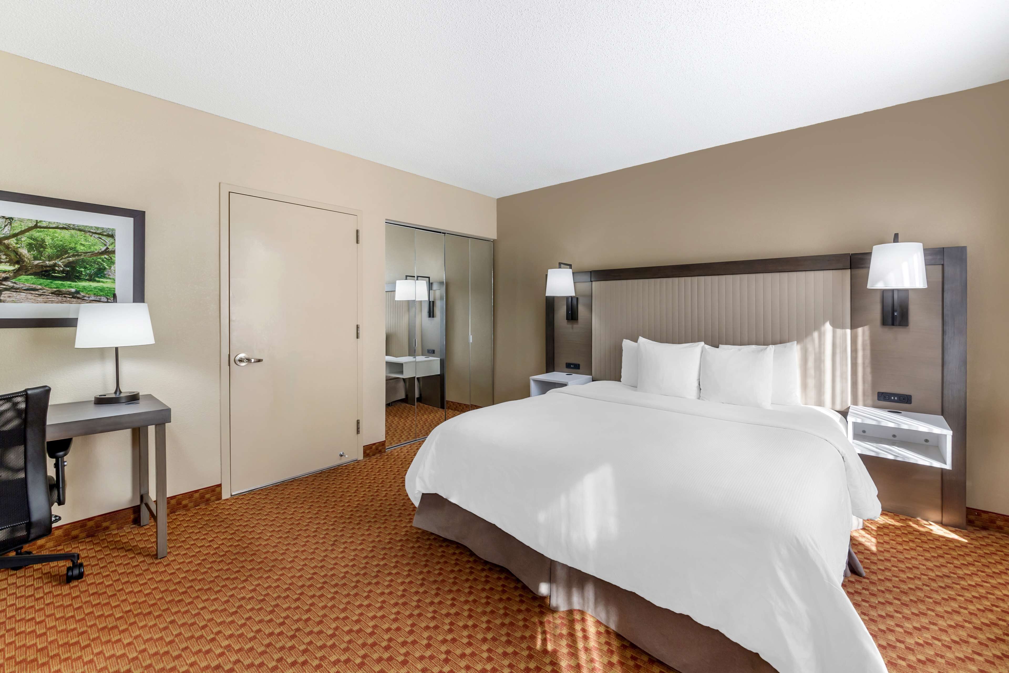 Ein Hotelzimmer mit einem großen Bett, einem Schreibtisch mit Stuhl, einem Nachttisch mit Lampe, einem Spiegel und einem Kleiderschrank.