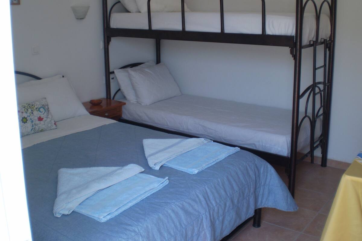 Habitación con literas, una litera inferior y una cama individual. Hay una mesita de noche pequeña con una lámpara y un almohadón.