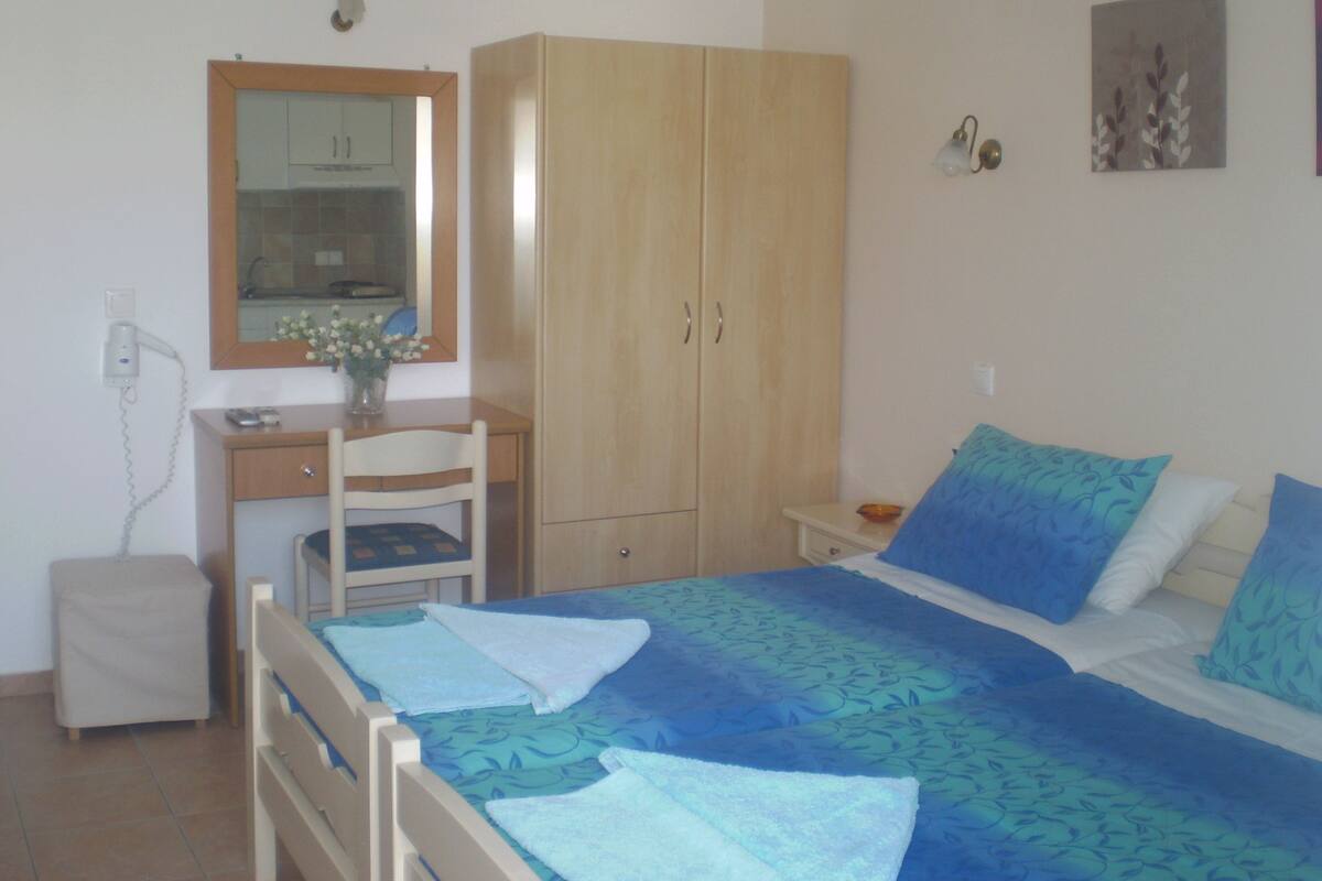 Un dormitorio con cama, un armario de madera, un escritorio con silla, una mesita de noche pequeña y un espejo montado en la pared.