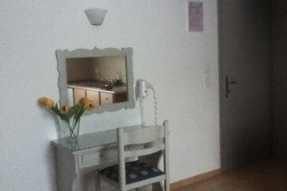 Una habitación con un escritorio pequeño blanco, un espejo y un florero con flores. Hay una puerta a la derecha y una lámpara en la pared.