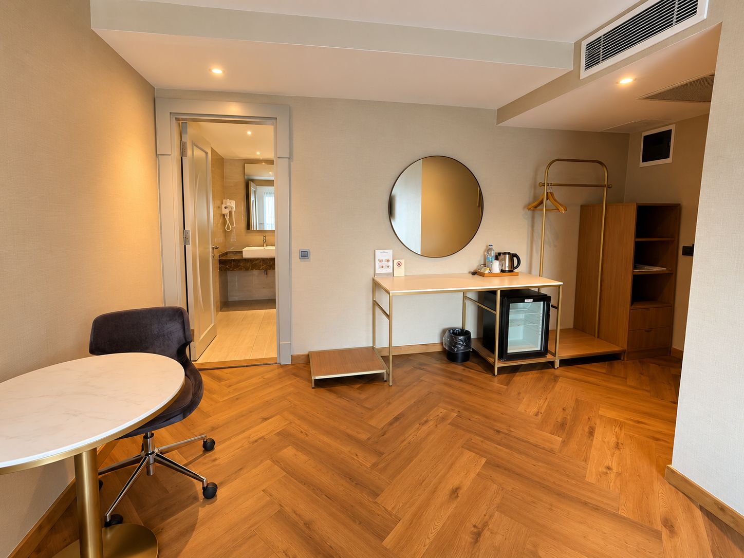 Hotellihuone, jossa on puulattia, pyöreä peili, pieni jääkaappi, työpöytä, tuoli ja vaatekaappi.