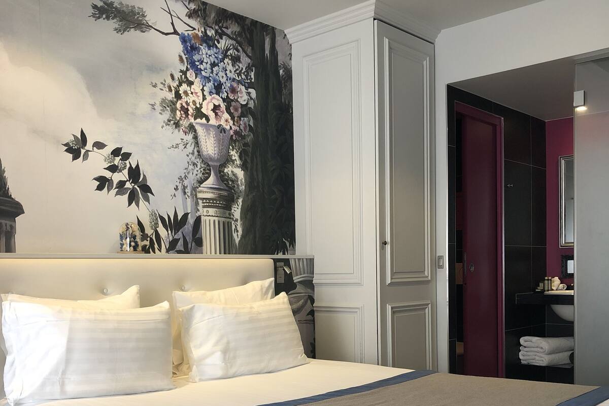 Un lit bien fait, avec du linge de lit blanc et un élément décoratif bleu, un grand vase rempli de fleurs sur le mur et une armoire avec un miroir.