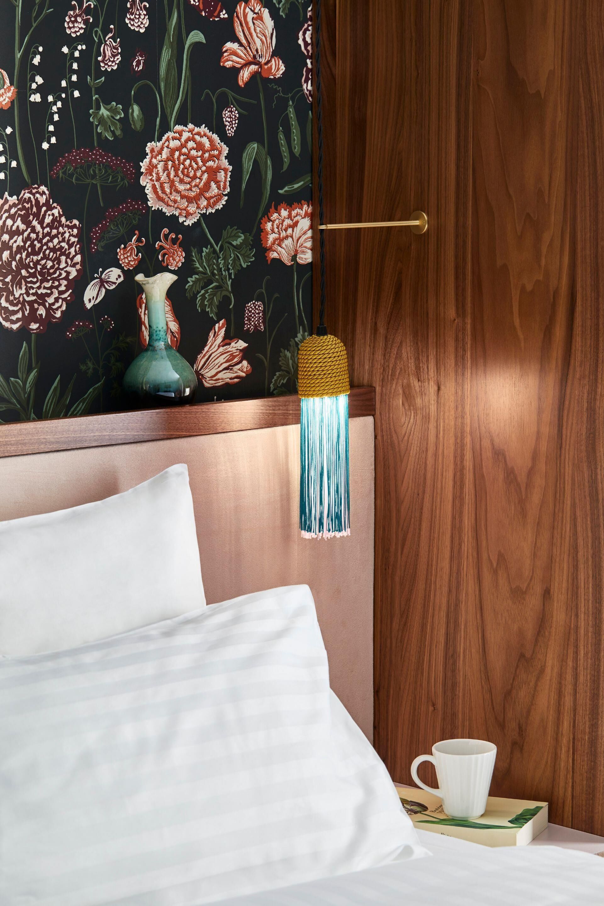 Una cama bien hecha con sábanas blancas, un florero verde aguamarina y una taza blanca sobre un libro, frente a un cabecero de madera y un papel tapiz floral.