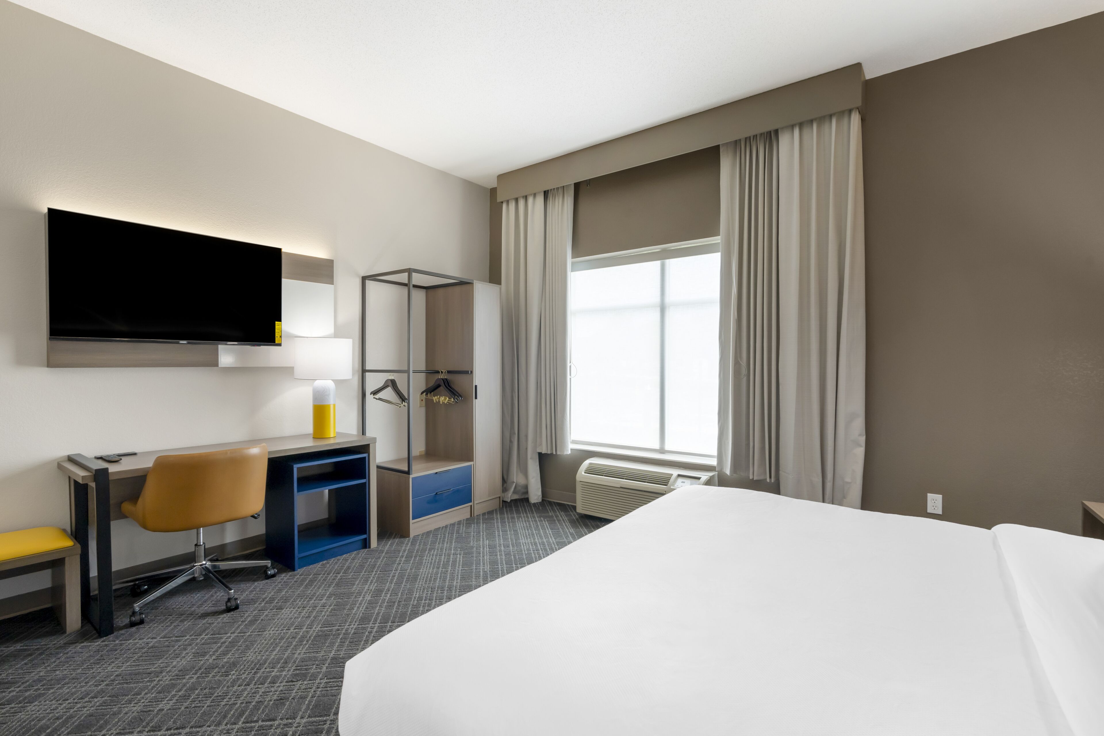 A hotel room with a bed, a desk with a chair, a TV mounted on the wall, and a window with curtains.