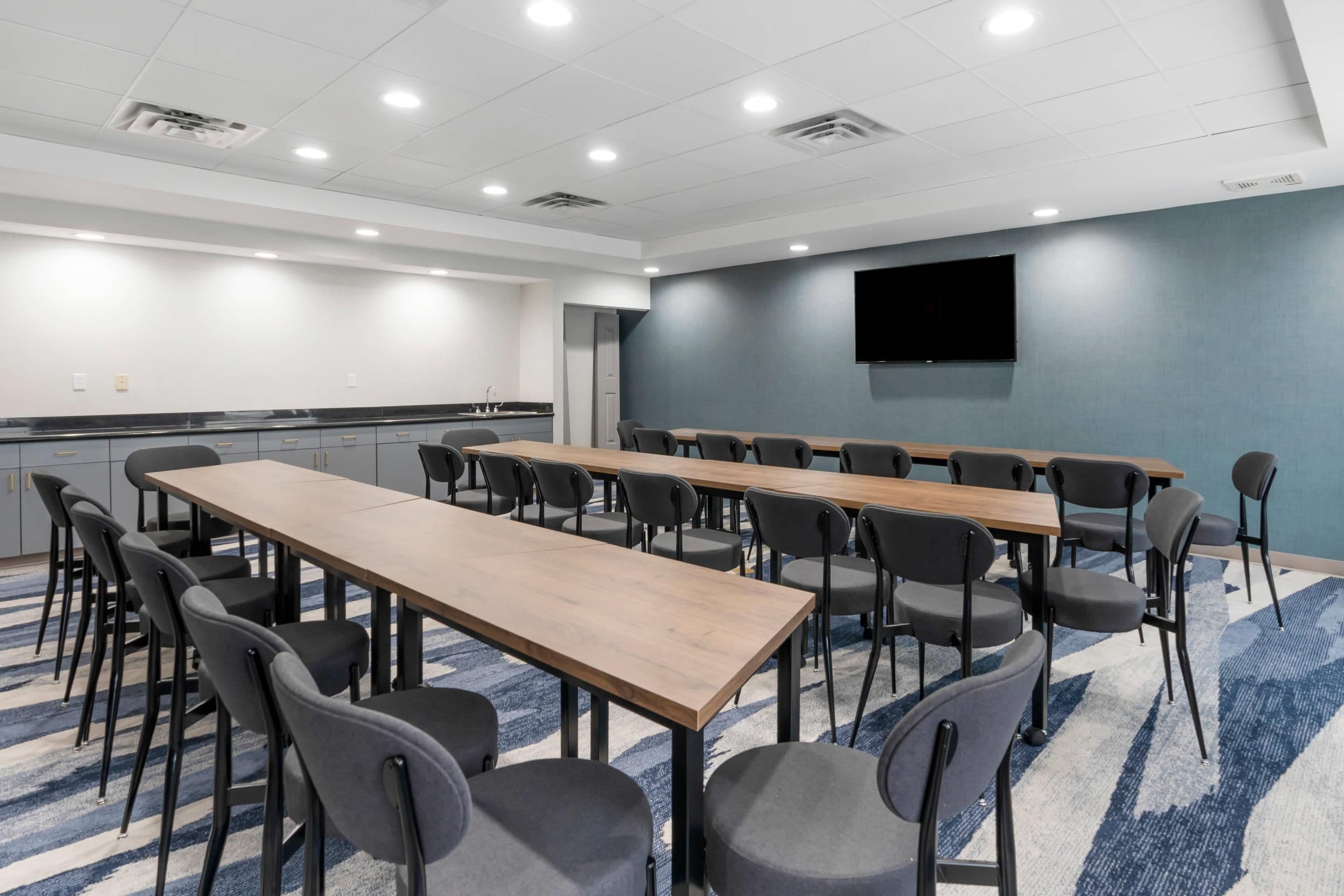 Un salón de conferencias con mesas largas y sillas, un televisor de pantalla plana y una zona de kitchenette.
