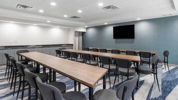 Un salón de conferencias con mesas largas y sillas, un televisor de pantalla plana y una zona de kitchenette.