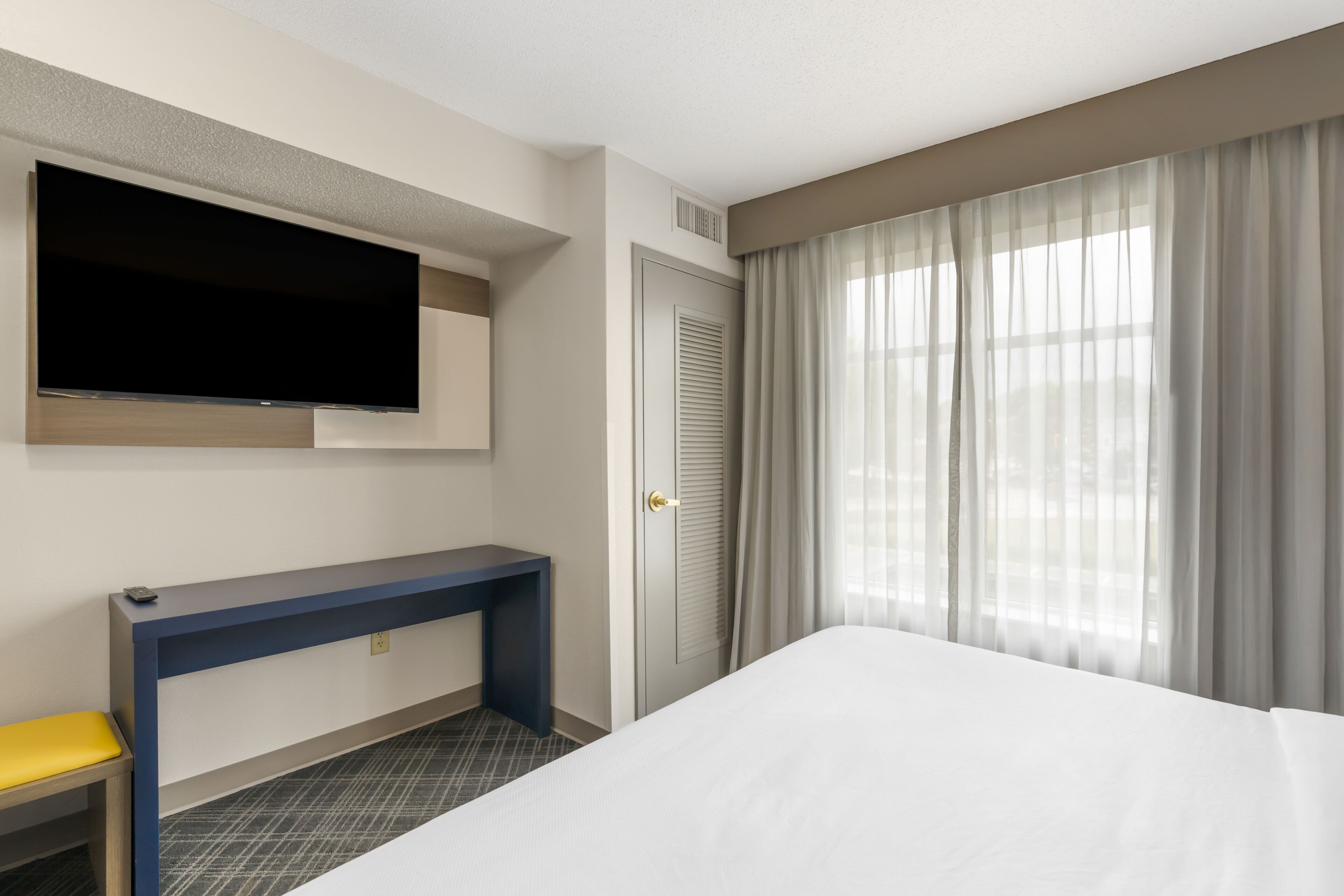Habitación de hotel con cama, escritorio, televisor montado en la pared, puerta con contraventanas y ventana con cortinas.