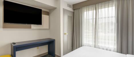 Habitación de hotel con cama, escritorio, televisor montado en la pared, puerta con contraventanas y ventana con cortinas.