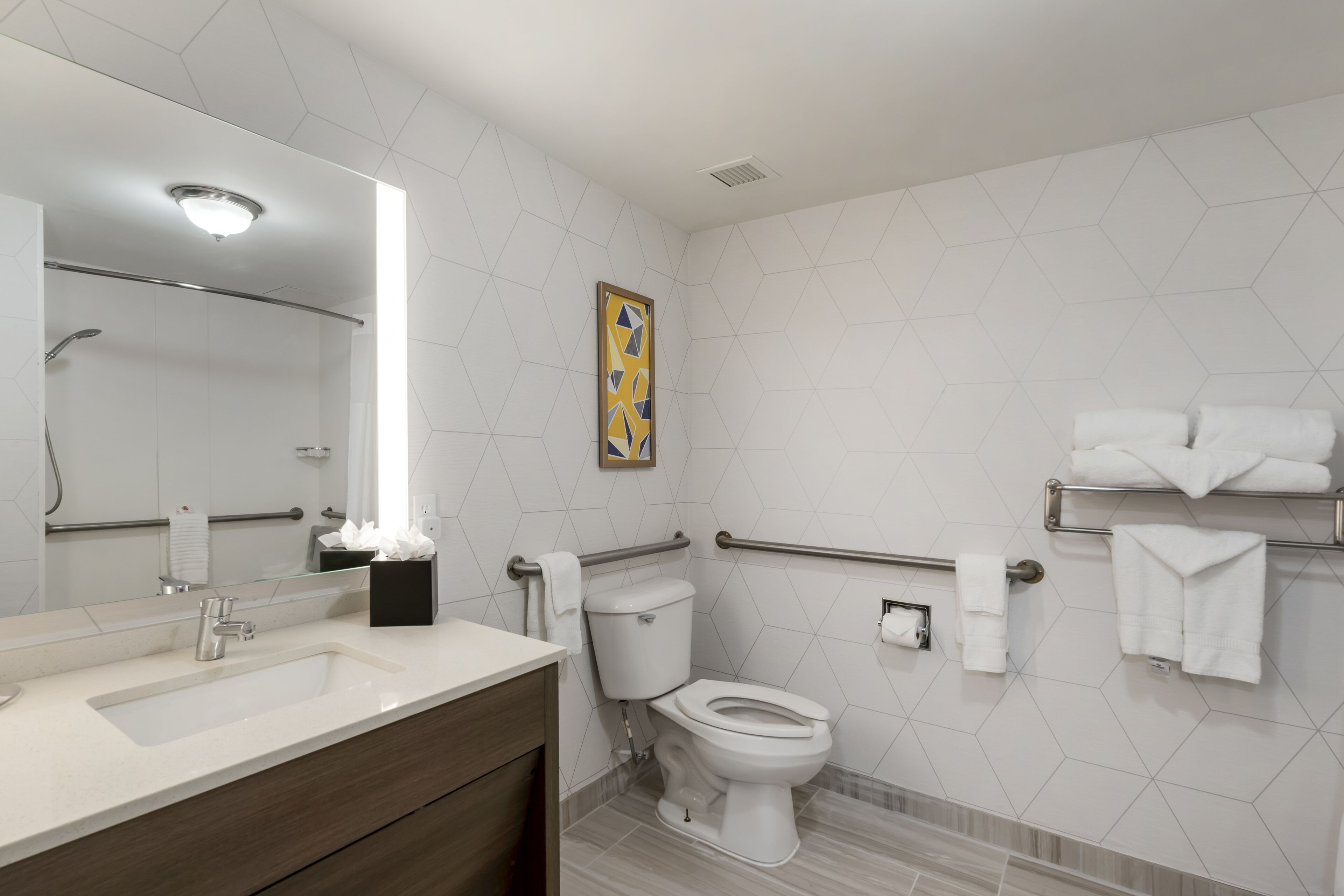 Un baño moderno con lavamanos, inodoro y ducha blancos. Hay barras de apoyo y un perchero.