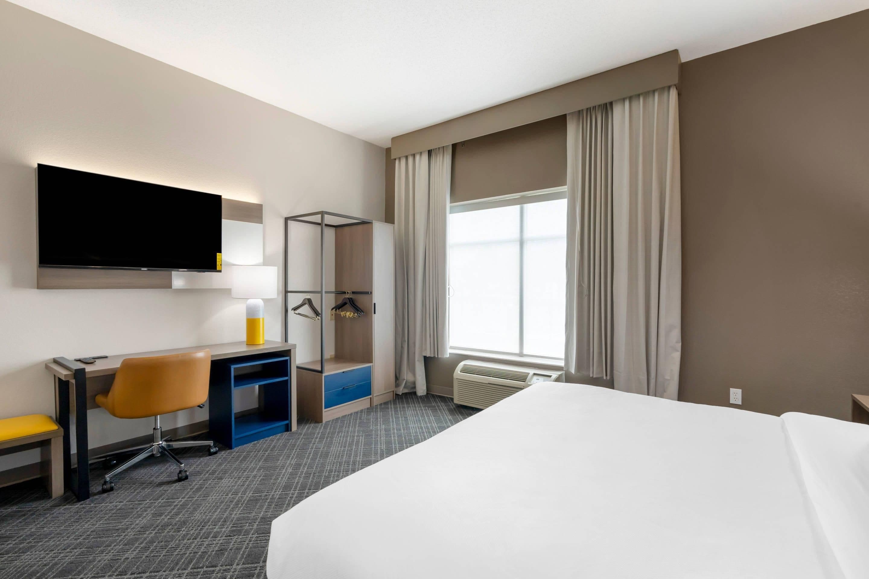 Une chambre d’hôtel comprenant un lit, un bureau avec une chaise, une télévision, une armoire et une fenêtre avec des rideaux.