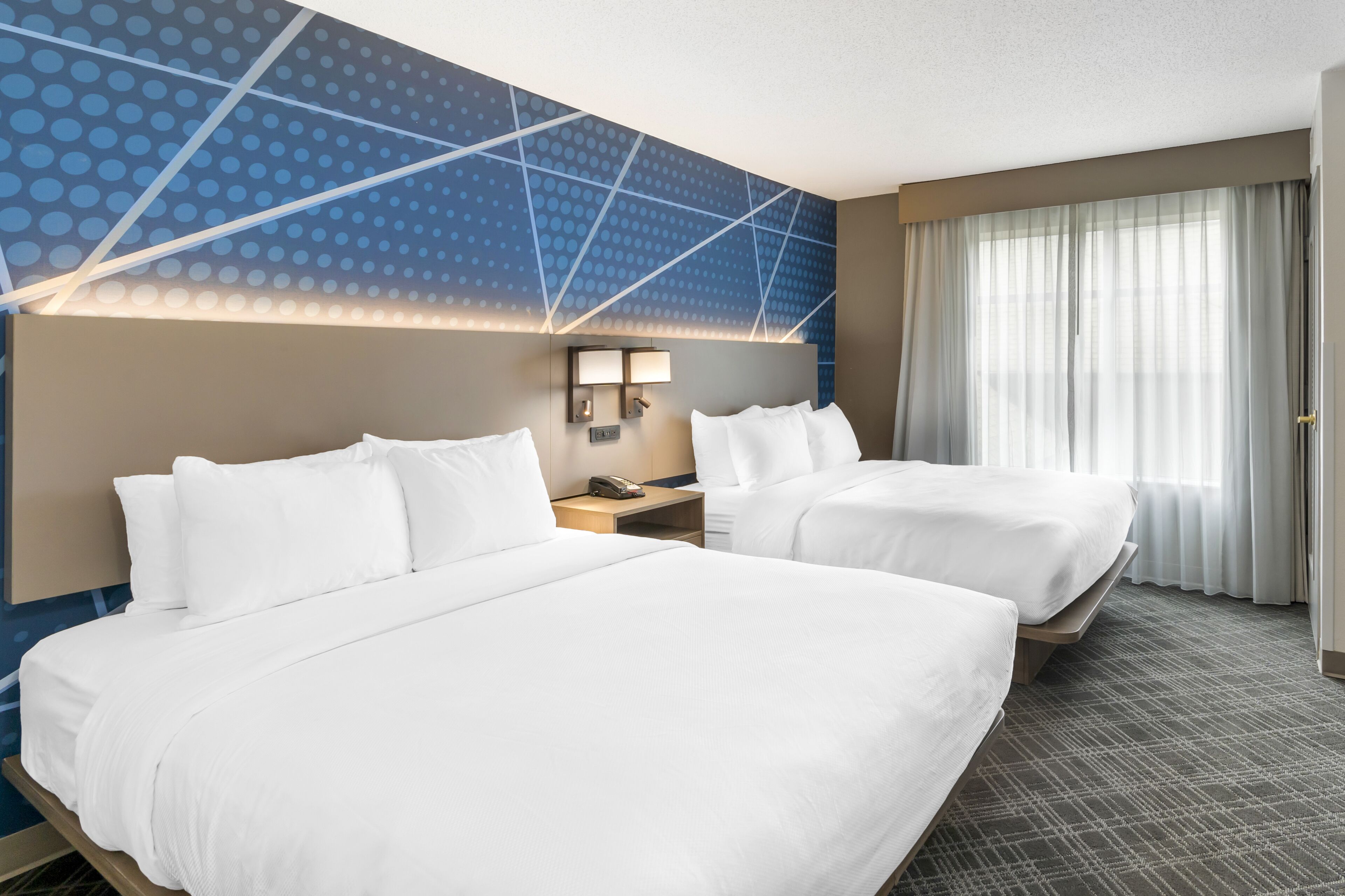 A hotel room with two beds, a nightstand with a phone, and a wall with a geometric pattern.