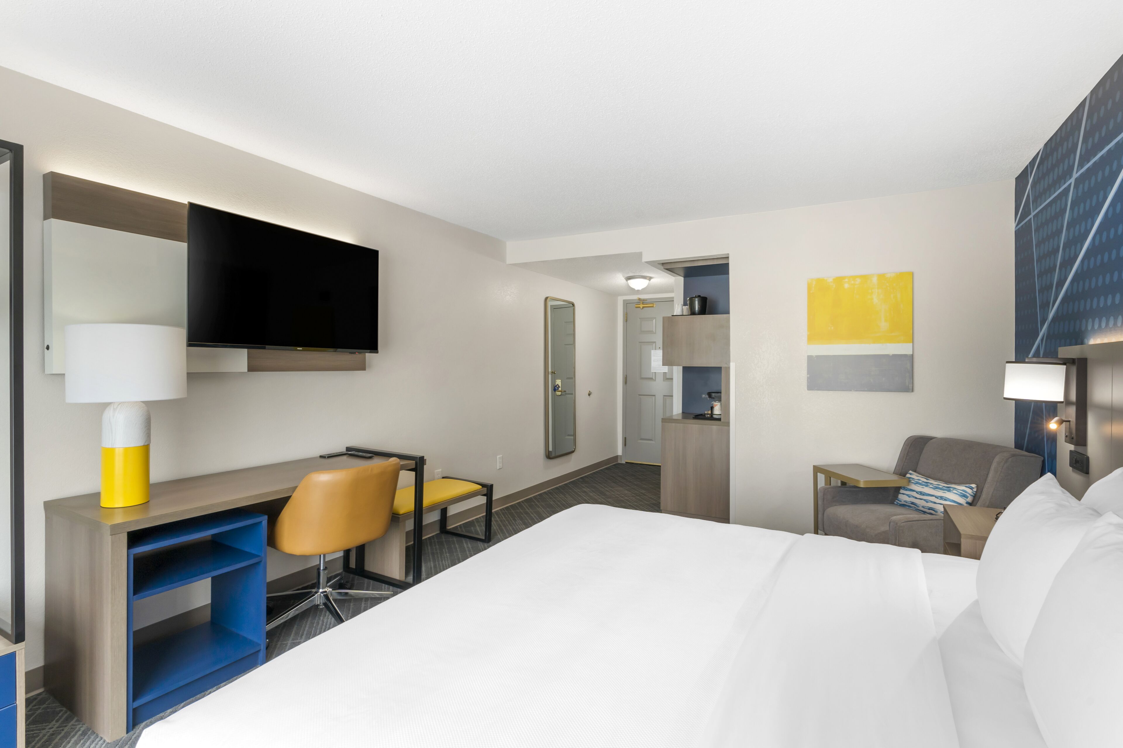 Habitación de hotel con cama, escritorio, silla, televisión y una pequeña mesa con lámpara.