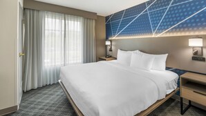 Hotellihuone, jossa on suuri sänky, yöpöytä, seinään kiinnitetty lamppu ja ikkuna, jossa on verhot.