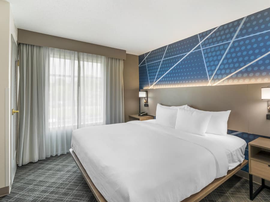 A hotel room with a large bed, a nightstand, a wall-mounted lamp, and a window with curtains.