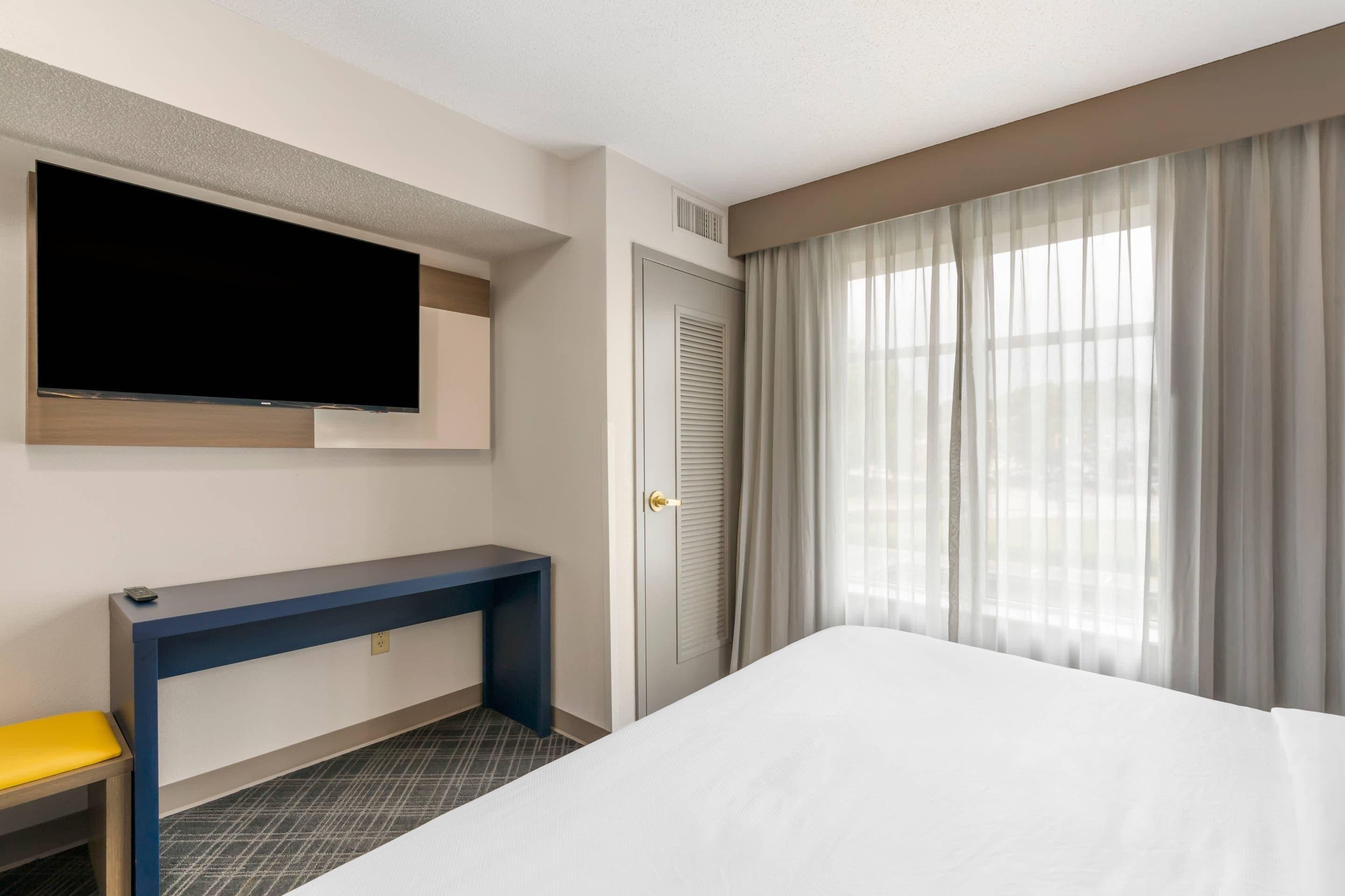 Habitación de hotel con cama, televisor montado en la pared, escritorio con silla y ventana con cortinas.